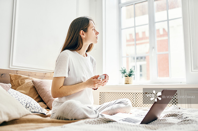 年轻女人手持白杯坐在床上，笔记本电脑旁思考图片素材