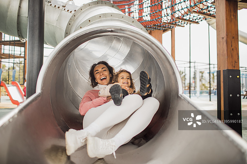 年轻快乐的母亲和女儿在游乐场的滑梯上玩耍图片素材