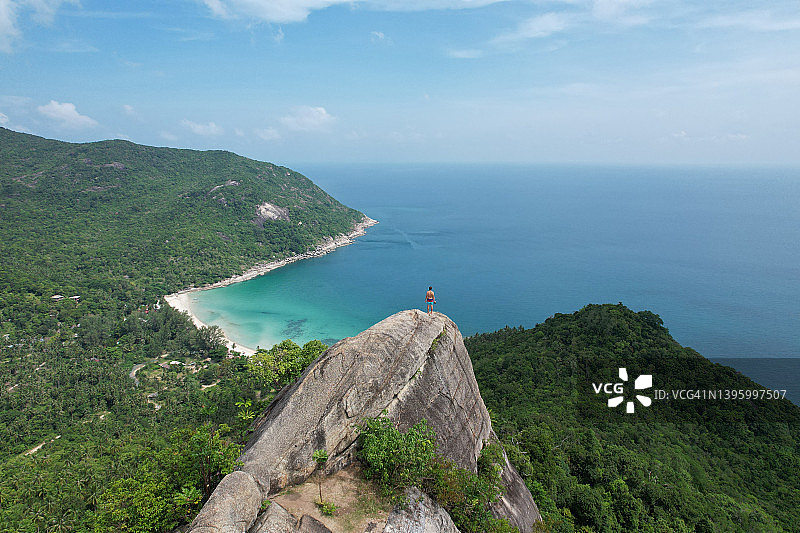 在岩石山顶上眺望大海和热带雨林景观的男人的无人机视角图片素材