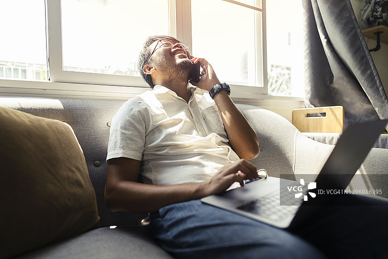 一位情绪积极的亚洲年轻自由职业者（数字游民）在家中使用手机和笔记本电脑工作，享受家庭生活图片素材