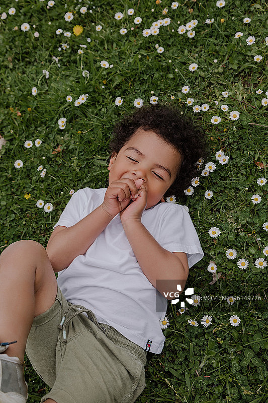 深色皮肤的男孩，留着爆炸头，躺在雏菊花的田野上笑着图片素材