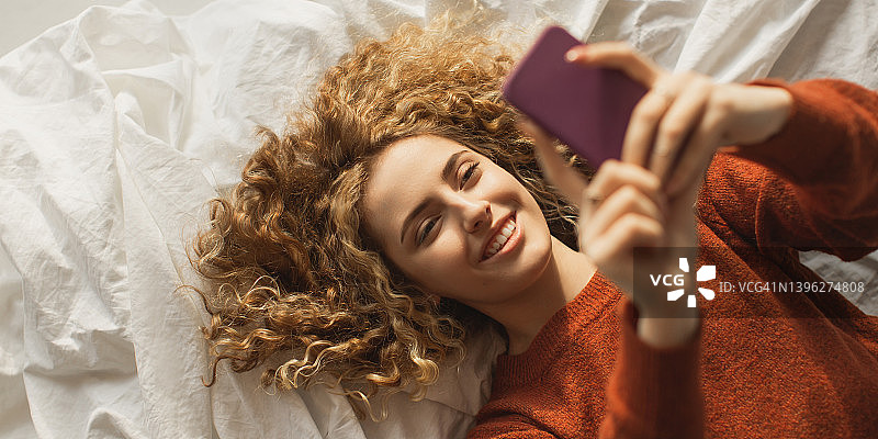 快乐的红发卷发女孩躺在床上俯视。身穿红色毛衣的女子正在看手机。喜悦。长长的卷发。网上银行。感受快乐。高架。在线交流。美丽的微笑图片素材