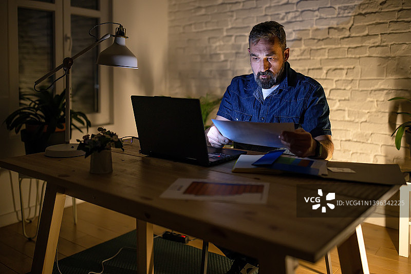 穿着休闲西装的中年男人坐在家庭办公室的桌子旁，在笔记本电脑上工作图片素材