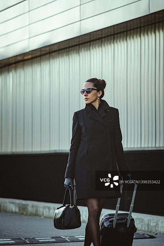 年轻优雅的女士在办公楼旁的便道上拉着小行李箱行走图片素材