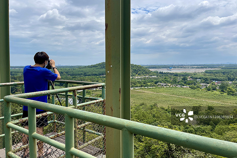 身穿蓝色T恤和短裤的男游客在山顶观景点拍摄罗勇府的风景，考博观景点图片素材