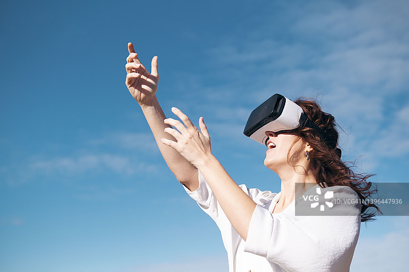 阳光明媚的日子里，站在天空前的年轻女子使用VR模拟器图片素材