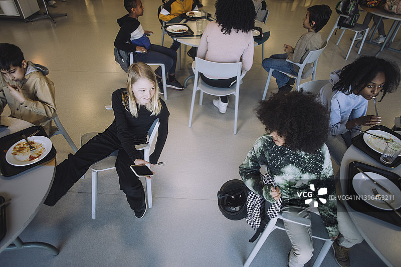 午休时，女孩在自助餐厅与坐在桌旁的朋友分享智慧手机图片素材