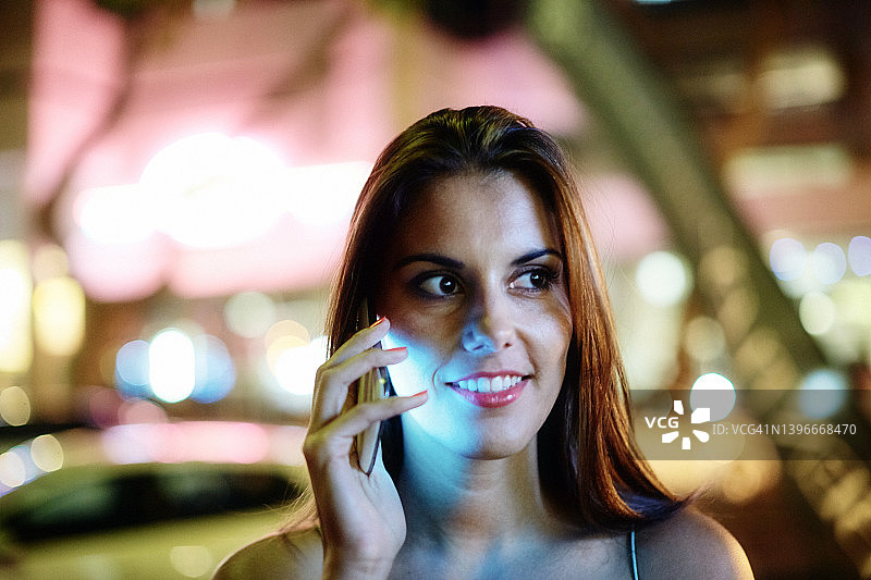 年轻女子在闪闪发光的夜总会外使用发光的手机时微笑图片素材