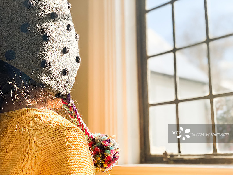 戴着绒球帽，穿着亮黄色毛衣的年轻混血女孩在晴天望着玻璃窗外图片素材
