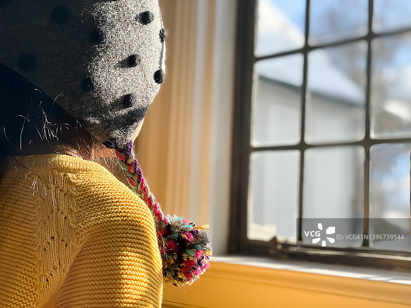 戴着绒球帽，穿着亮黄色毛衣的年轻混血女孩在晴天望着玻璃窗外图片素材