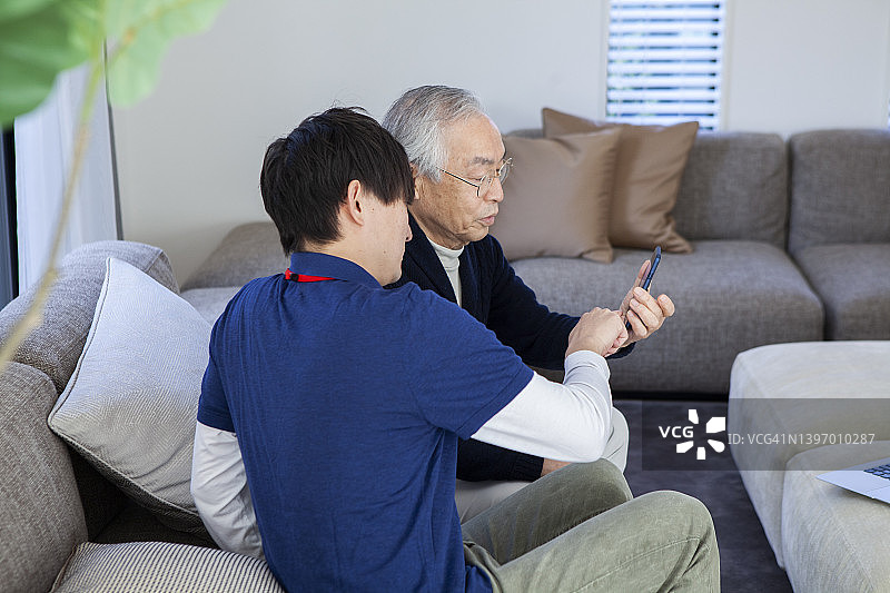 年长的男人在客厅里通过智能手机与家人交流，年轻的护理人员在一旁协助图片素材