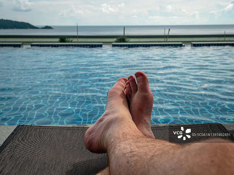 年轻白人男性在热带豪华酒店度假村的阳光下放松，海滩和游泳池边的日光躺椅。健康福祉、休息、自由、假期、度假和旅行的概念图片素材