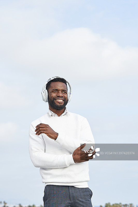 在晴朗的天空下戴着耳机听音乐的快乐的非裔美国男人图片素材