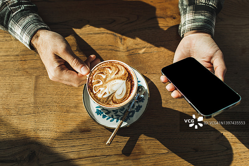 俯视：男人手持咖啡杯和手机，木桌背景图片素材