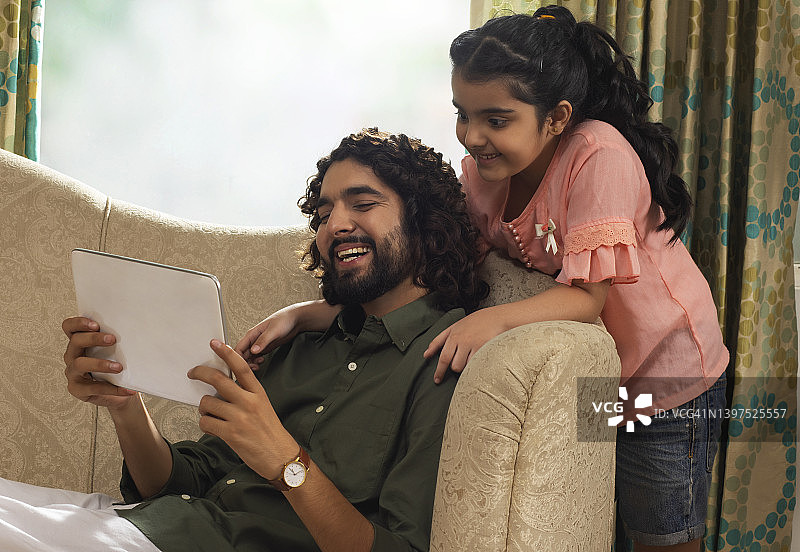 父亲在沙发上看平板电脑，女儿站在沙发后面图片素材