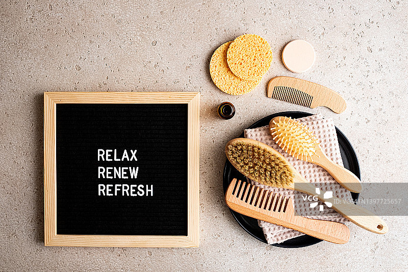 黑色托盘上的环保型头发和身体护理用品套装（发刷、清洁海绵、面部精华液、木制身体刷、棉毛巾），以及带有文字“放松、焕新、清爽”的留言板，混凝土背景，俯视图。家庭水疗概念。零浪费图片素材