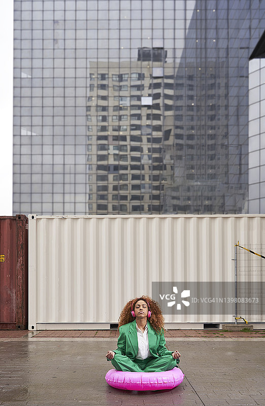 年轻女商人戴无线耳机听音乐，坐在粉色充气圈上冥想图片素材