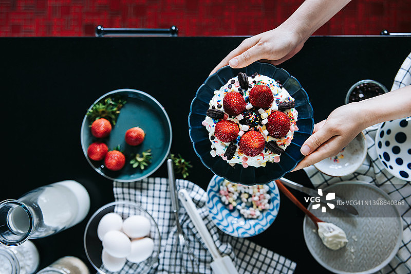 女人手持自制海绵奶油蛋糕，用草莓、棉花糖、巧克力和糖屑装饰。厨房台面上摆放着各种烘焙用品。在家烘焙。生日和庆祝概念图片素材
