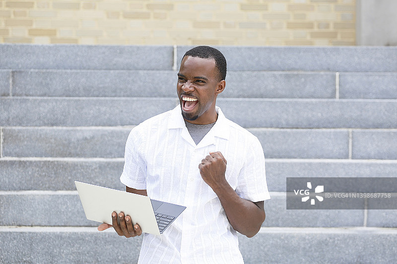 一名留着山羊胡的非裔美国千禧一代男子手持笔记本电脑，面露兴奋之色，身穿白色衬衫图片素材
