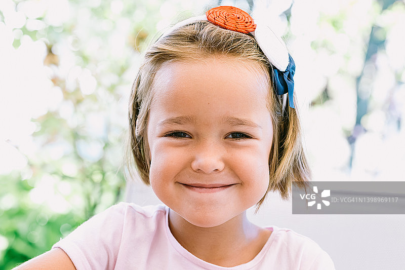 微笑的金发小女孩肖像，戴蓝红白装饰的白色发带，背景是花园图片素材