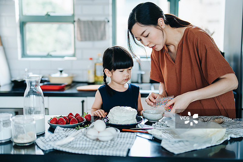 年轻的亚洲母亲和小女儿在厨房里烤蛋糕，并一起用草莓、搅打奶油和糖屑装饰。家庭烘焙。DIY。生日和庆祝概念图片素材
