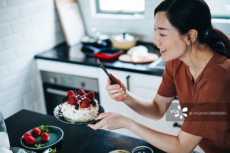 年轻的亚洲女性在家烤蛋糕，用智能手机拍摄她新鲜的自制蛋糕。厨房柜台上摆放着各种烘焙用品。在家烘焙。DIY。生日和庆祝概念图片素材