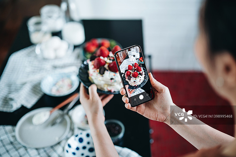 年轻亚洲女性在家烘焙蛋糕，用智能手机拍摄自制蛋糕肩部视角，厨房台面有各种烘焙用品，生日和庆祝概念图片素材