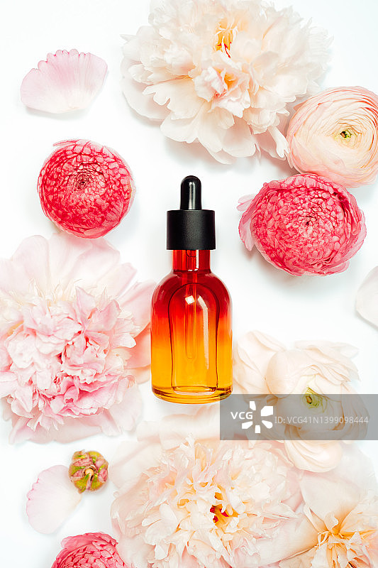 白色背景上带有柔和粉色牡丹和毛茛的橙色玻璃瓶面部精华液特写图片素材