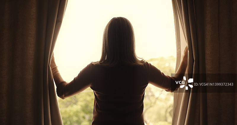 一个成熟的女人在家里放松的早晨打开她卧室的窗帘。后视图的年老妇女的剪影看窗外，开始一天图片素材