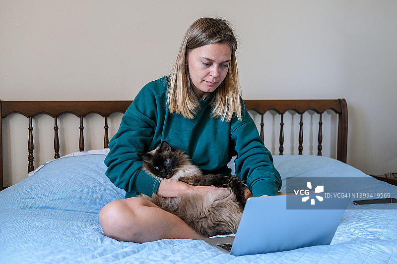 年轻女人在家中床上用笔记本电脑工作，抱着猫，看剧或社交，远程教育或工作图片素材
