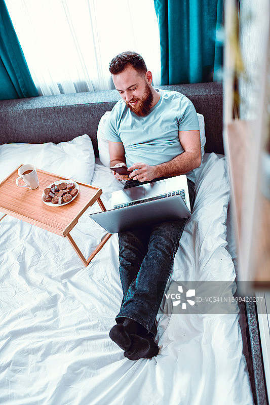 胡子男在床上放松，享用零食、咖啡，使用笔记本电脑和社交媒体图片素材