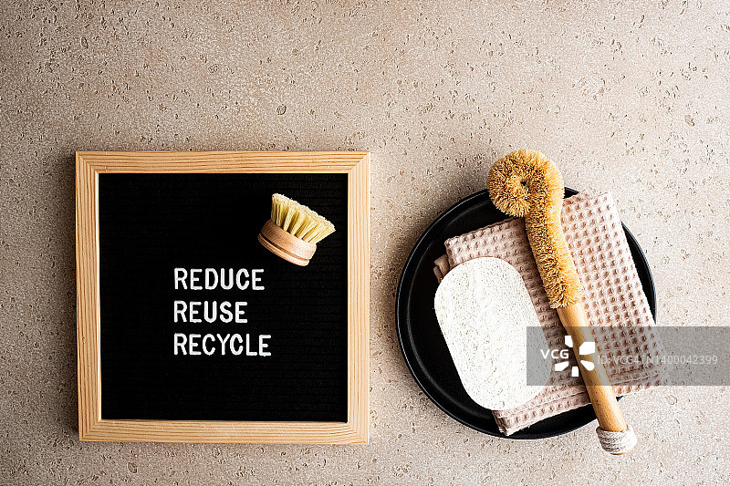 黑色托盘上的环保厨房清洁用品（木制洗碗刷、棉布、丝瓜海绵）以及混凝土背景上的“减少、再利用、回收”字样的信板，俯视图。清洁设备。零浪费理念。图片素材