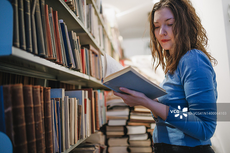 美丽的女大学生站在图书馆的书架旁，手捧着打开的书图片素材