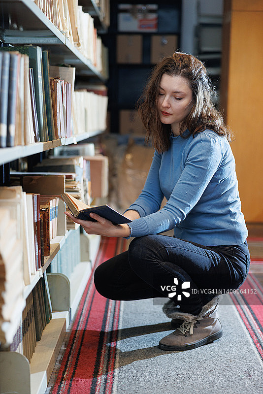 美丽的女人蹲在大学图书馆的书架旁，手捧一本打开的书阅读图片素材