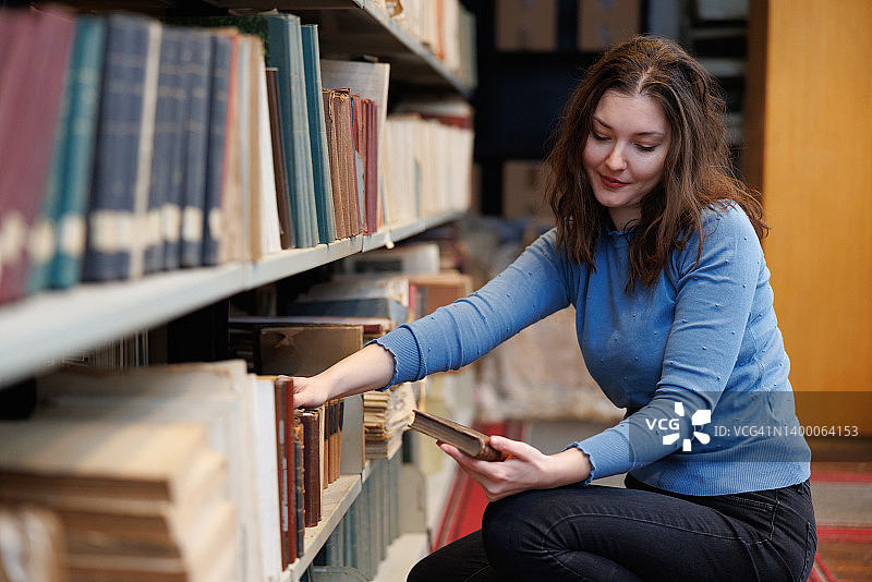 美丽的女人蹲在大学图书馆的书架旁，拿着一本书看着封面图片素材