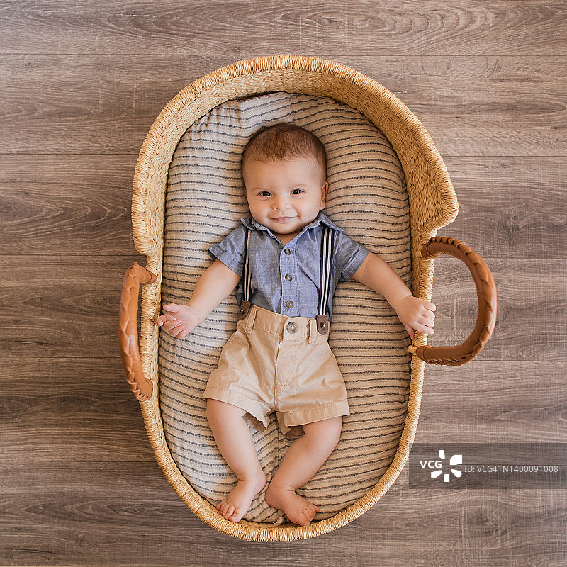 一个22周大的男婴穿着蓝色背带裤套装，躺在海草摩西篮里舒适的奶油色条纹棉毯上图片素材