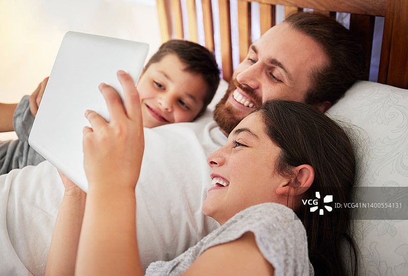 快乐的白人父亲和两个孩子在床上用平板电脑一起看电影、阅读或玩游戏图片素材