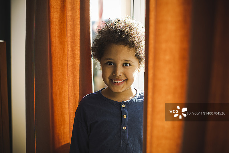 微笑的男孩站在家里的窗帘后面的肖像图片素材