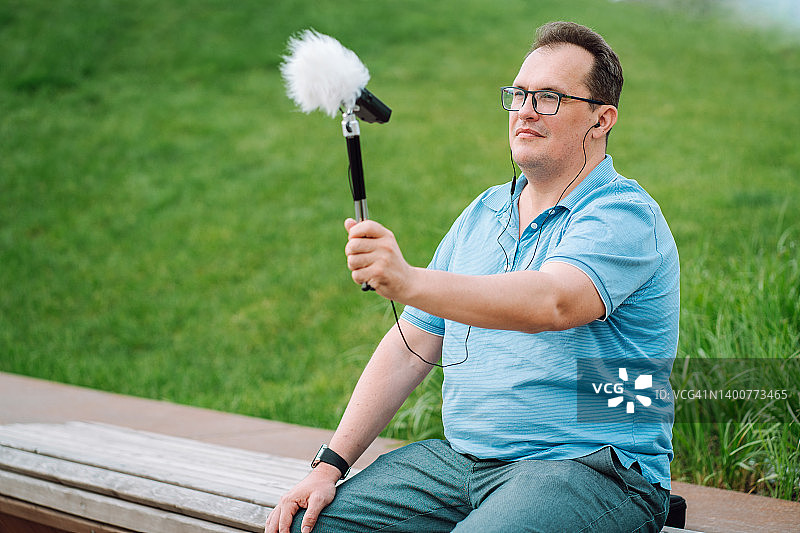 戴眼镜的深色头发中年男人坐在公园长椅上，用麦克风和仪器测量噪音图片素材