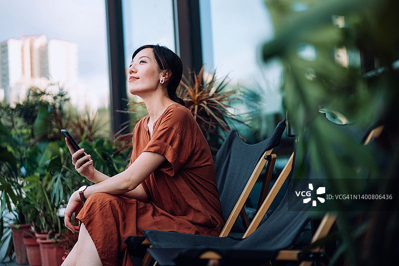 笑容甜美的年轻亚洲女人在公寓阳台上放松的早晨，坐在躺椅上使用智能手机，周围环绕着漂亮的室内植物。发短信、休闲和生活方式。日常生活中的科技图片素材