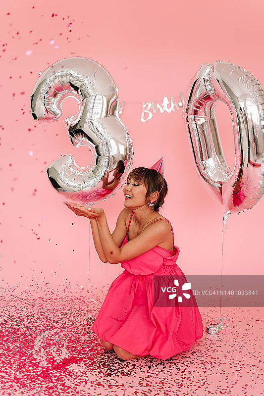 年轻女子身穿粉色迷你裙，在影棚粉色背景的彩带气球中庆祝三十岁生日图片素材