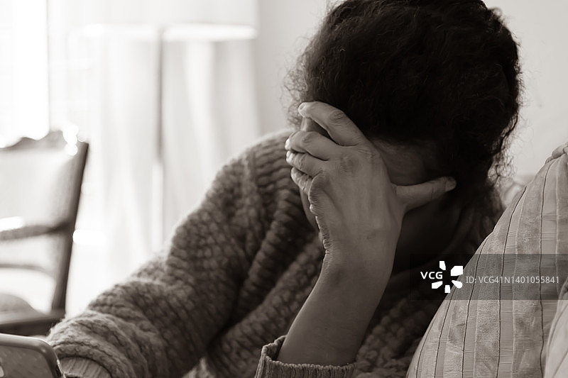 美国新的精神抑郁症：沮丧的黑人妇女坐在沙发上，手扶着头，拿着智能手机图片素材