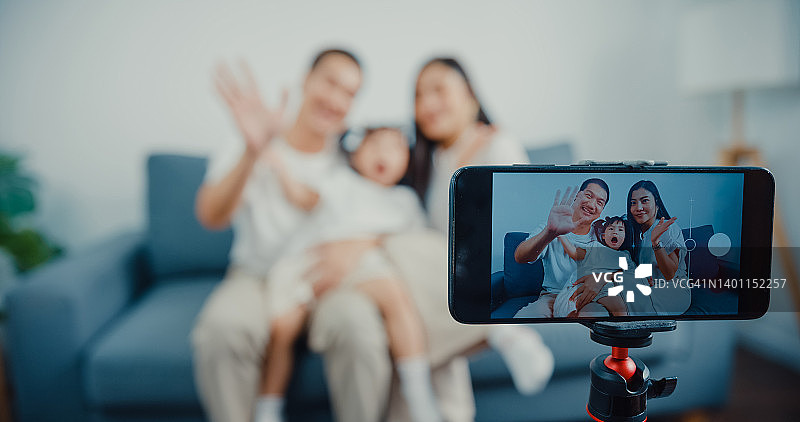 年轻的亚洲父母坐在沙发上，在家中使用智能手机通过摄像头打招呼图片素材