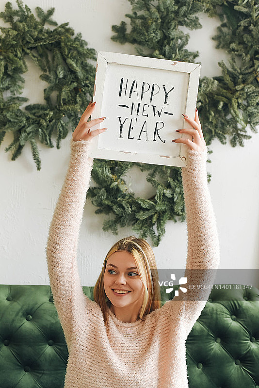 在新年的背景下手持标牌的微笑女孩。新年快乐标牌。圣诞节背景。金发女孩。蓬松的毛衣。新年花环。图片素材