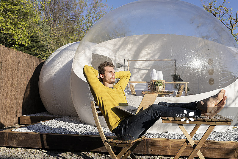 坐在透明圆顶酒店前椅子上，手提笔记本电脑的沉思的年轻商人图片素材