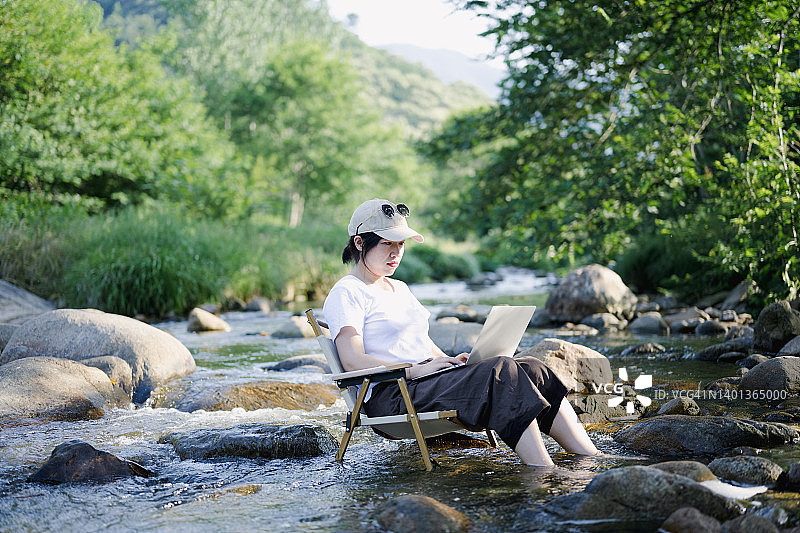 一位戴帽子的亚洲年轻女性坐在溪流中的椅子上用笔记本电脑工作图片素材