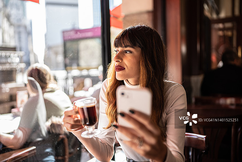 在咖啡馆里，沉思的年轻女人望着窗外，手持手机和咖啡杯图片素材