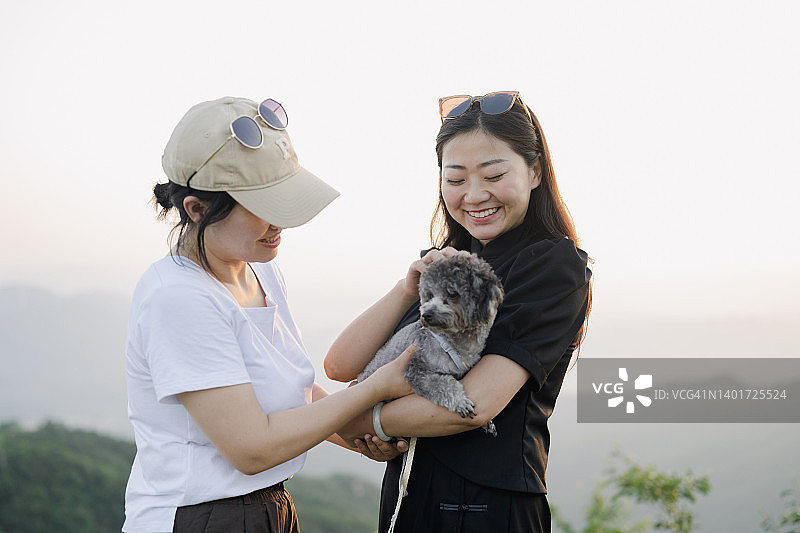 两位年轻的亚洲女性拥抱小狗，背景是晴朗的天空和山脉图片素材