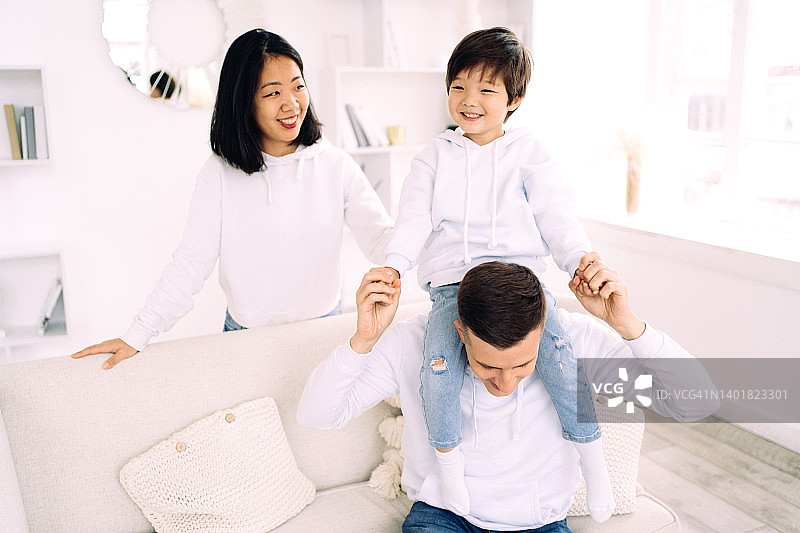 年轻的多种族家庭在客厅里休息，父亲让儿子坐在肩膀上，母站在他们身后，家庭生活概念图片素材