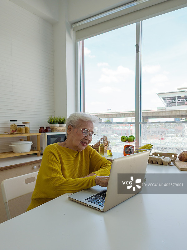 亚洲老年妇女早晨在厨房使用笔记本电脑进行电子学习图片素材